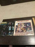 Two Rolling Stones albums with sleeves on a dark wooden surface, showing front and back covers with clear visibility of album titles and artwork.