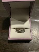 Front-facing image of silver ring with row of clear stones displayed in pink and white ring box on dark wooden surface.