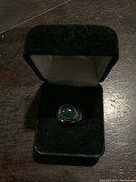 Silver ring with green round cabochon stone set in silver band, displayed in black velvet ring box on a wooden surface.