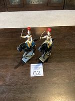 Two metal toy soldiers on black horses with red plume helmets and bows, shown from front and angled side view on wooden surface.