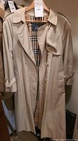 Full front view of beige trench coat hanging, showing buttons, belt and overall style