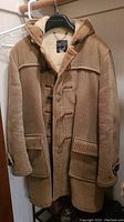 Full view of tan corduroy duffle coat hanging on hanger showing front with toggles, pockets, hood and cuff leather straps
