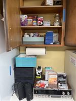 Wide view showing wooden shelving with multiple light bulb packages, stereo system unit with knobs visible, black stereo speakers, Sony radio, and CDs stacked on the stereo unit.