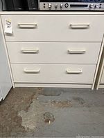 White storage cabinet with three horizontal drawers, white handles, seen from the front on a concrete floor.