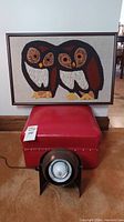 Full view showing framed fabric owls resting on the red vinyl ottoman foot stool and brown swivel ball lamp in front, on carpet.