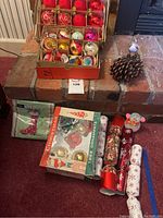 Photo of vintage box with 16 assorted round glass Christmas ornaments in original packaging, a pinecone, an acorn decoration with bird, pack of Christmas napkins, snowman figure, and six Christmas crackers arranged on carpet and brick hearth.