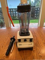 Vita-Mix blender fully assembled with pitcher, lid, and tamper placed beside the base on wooden table under window light.