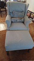 Front view of beige upholstered wingback chair with two pillows and matching ottoman in carpeted living room.