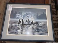 Framed numbered print 'Whale Song' showing orca whales swimming and splashing in water with forested background, black frame and white matting
