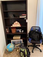 Full view of tall dark wood bookshelf with five shelves containing various books and accessories. Next to it is a blue office chair with black armrests and a mesh back. Floor items include a globe on a brass stand, a brass table lamp, and a green and black emergency backpack.