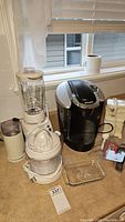 Overview photo of Keurig coffee maker, blender, juicer, can opener, and other small kitchen items on countertop by window.