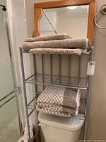Photo showing grey metal bathroom storage rack placed over the toilet, holding folded towels on top and middle shelves beneath a light wooden framed mirror