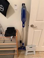 Hoover stick vacuum cleaner standing against a white wall next to a wooden shoe rack, black heater/fan and fan on shoe rack.