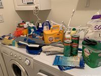 Photo showing multiple cleaning product bottles and tools on top of washing machine and dryer, including detergents, cleaners, and scrub tools.