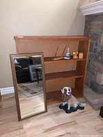 Wooden bookshelf with two shelves, wooden frame mirror leaning against it, and ceramic dog figurine on the floor in front.