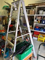 Full view of the aluminum extension ladder, green tarp folded inside plastic container beneath it, with wooden bench beneath the container.