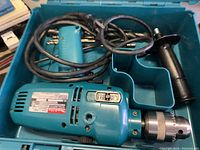 Makita HP310 hammer drill with side handle and cord in teal carrying case, with drill bits included.