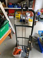 Full view of hand truck standing upright with several heavy duty clamps hanging from the top and spirit level resting against the wheels. Plastic container at base containing the orange electric jig saw with power cord wrapped.