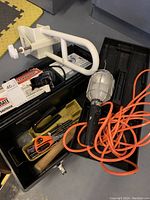 Photo showing the open black toolbox with various tools including a handheld work light with orange power cord, white clamp, hammer, scissors, drill bits, and screwdriver inside.