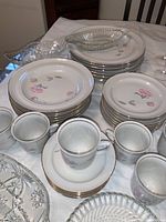 Photo shows multiple stacked plates in several sizes with pink flower and gold trim, tea cups and saucers, and crystal dishware pieces in background.