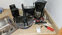 Kitchen starter set items arranged on a beige countertop next to a sink and backsplash with white tiles, showing multiple small appliances and utensils with a numbered lot tag.