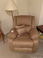 Full view of a light tan fabric recliner chair with plush armrests and a striped accent pillow. Next to the chair is a tall brass floor lamp with a beige lampshade.