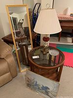Mid century round wooden table with glass top, brass table lamp, gold framed mirror standing behind sofa, and framed blue bird artwork leaning at table base.