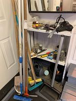 Image shows various tools including a large shovel with wooden handle, a broom with turquoise bristles, a rubber mallet, and hand tools on metal shelving. Also visible is a black corded power tool and containers of small parts or hardware.