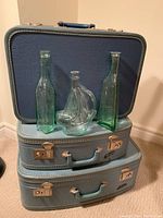 Photo showing the stack of three vintage blue suitcases with three glass decorative bottles placed in front: two green rectangular bottles and one clear ship-shaped bottle.