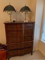 Full view of vintage Bassett tall boy chest of drawers with two brass touch lamps on top, set against a beige wall near a window