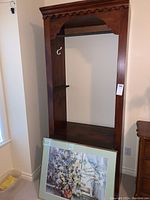 Wooden hanging storage cabinet with a clothing bar inside and scalloped trim, paired with framed floral artwork by Brent Heighton leaning against the bottom shelf.