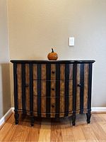Front view of a black and gold striped cabinet with three center drawers and side doors closed. Wooden floor and light wall background.