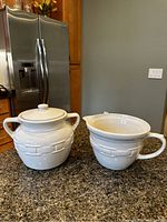 White Longaberger jar with lid and matching cup or bowl on kitchen counter, stainless steel refrigerator in background.