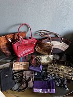 Full view of various purses and bags arranged on a table against a gray wall including woven leather tote, red leather handbag, straw and leather shoulder bag, and multiple smaller clutches and pouches.
