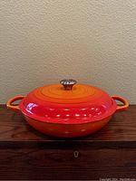 Front view of orange-red enameled Le Creuset casserole dish with lid on wooden surface