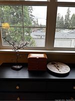 Three decor items arranged on a black tabletop in front of a window including a metal tree jewelry holder, wooden box, and sunflower clock.