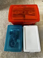 Three plastic storage bins seen from above; orange bin on top filled with cards, medium blue and white bins below containing electronics and cables