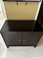 Full front view of Mark Thomas cherry cabinet with two doors and silver handles on light wooden floor under TV.