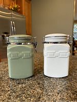 Photo showing front view of two Longaberger spice jars, one green and one white, placed on kitchen countertop.