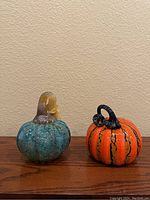 Blue-green and orange glass pumpkins displayed side by side on wooden surface against plain wall, showing front and side views in three photos.