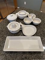 Several white Longaberger pottery pieces including covered container, nested bowls, double bowl with handle, citrus juicer, and rectangular platter on granite countertop