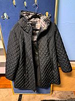 Full view of the black quilted coat hanging on a hanger showing the front with visible hood and zippered pockets.