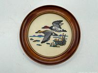Overall front view of round wooden framed cross-stitch wall art depicting two flying ducks over water.