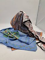 Two silk scarves displayed, one blue with white polka dots and floral pattern, the other white with black and red abstract florals