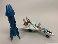 Photo showing the F-14A Navy jet fighter toy plane with detailed paint markings alongside the blue mechanical spaceship rocket coin bank standing upright.