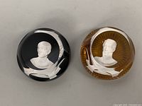 Two round Baccarat paperweights displayed side by side. Left one black with Julius Caesar bust, right one amber with Simon Bolivar bust.