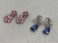 Two pairs of 925 silver drop earrings shown on white background. One pair pink floral crystal clusters, the other blue pear-shaped crystals with clear stones.