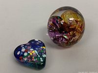 Two Robert Held blown art glass paperweights shown side by side, one round with amber and purple swirls, the other heart-shaped with multicolored floral design on dark blue background.