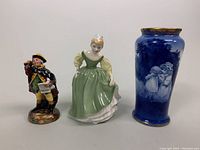 Photo of all three Royal Doulton items: Fair Maiden figurine, Town Crier figurine, and cobalt blue vase grouped together showing details, colors, and scale.