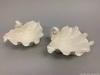 Two white porcelain bowls shaped like clam shells with detailed shell and sea creature feet from a top side angle